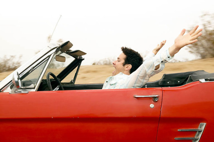 Fotografie muže, který jede v červeném kabrioletu se zvednutýma rukama