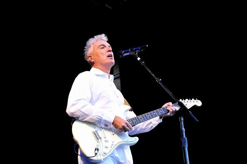 Muž v bílém obleku s bílou kytarou zpívá na pódiu